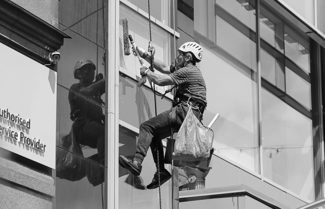 services-01 Black and white from below of man steeplejack in protective helmet with professional equipment cleaning glass wall of modern tower